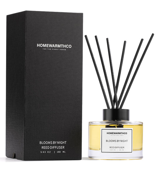 A reed diffuser set, with a clear bottle containing oil and black reeds, placed next to its black packaging box with the brand 'HOMEWARMTHCO' visible on it.