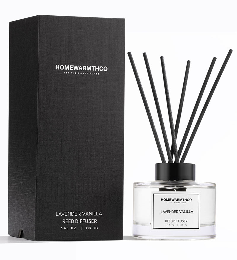 A luxury reed diffuser from HomeWarmthCo with black reeds in a clear glass bottle, labeled Lavender Vanilla. The product is next to its black box packaging with the same branding and product name.
