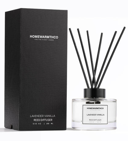 A luxury reed diffuser from HomeWarmthCo with black reeds in a clear glass bottle, labeled Lavender Vanilla. The product is next to its black box packaging with the same branding and product name.
