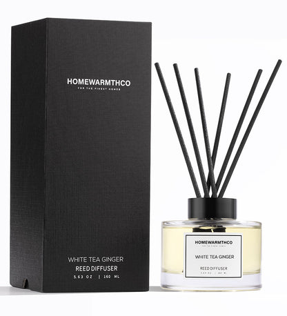 A luxury reed diffuser by HomeWarmthCo with black reeds in a clear bottle, labeled White Tea Ginger. The product is displayed alongside its black packaging box.